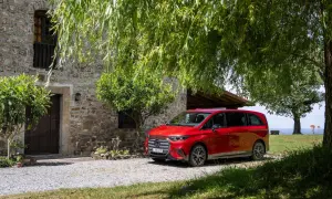 Die neue V-Klasse in Rot parkt vor einem Steingebäude unter Bäumen.