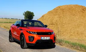 Ein leuchtend orangefarbener Range Rover Evoque Cabrio steht auf einem Feldweg neben einem riesigen Strohballen.