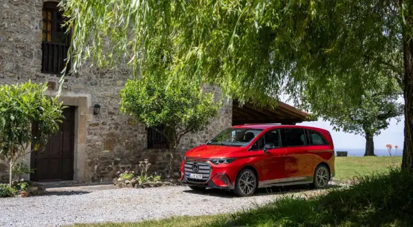 Die neue V-Klasse in Rot parkt vor einem Steingebäude unter Bäumen.
