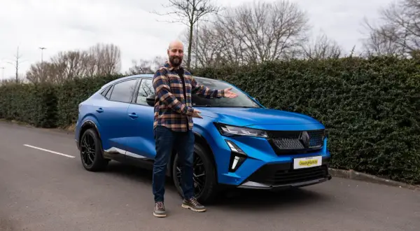 Ein Mann präsentiert stolz den neuen, blauen Renault Rafale vor einer Hecke.