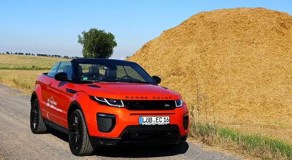 Ein leuchtend orangefarbener Range Rover Evoque Cabrio steht auf einem Feldweg neben einem riesigen Strohballen.