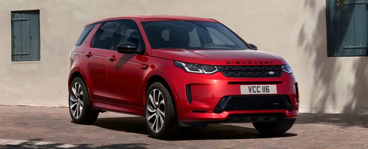 Roter Range Rover Discovery Sport, ein **range rover approved** Fahrzeug, parkt vor einer hellen Wand mit grünen Fensterläden.