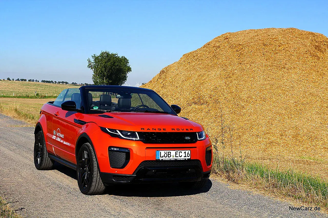 Ein leuchtend orangefarbener Range Rover Evoque Cabrio steht auf einem Feldweg neben einem riesigen Strohballen.