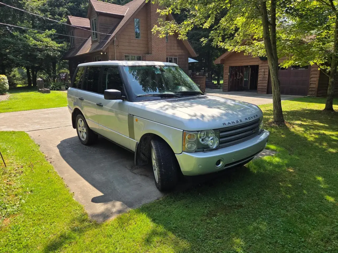 Silberner Range Rover Sport parkt vor einem Holzhaus. Möglicherweise gibt es Probleme mit der Luftfederung, da das Fahrzeug etwas tief steht.