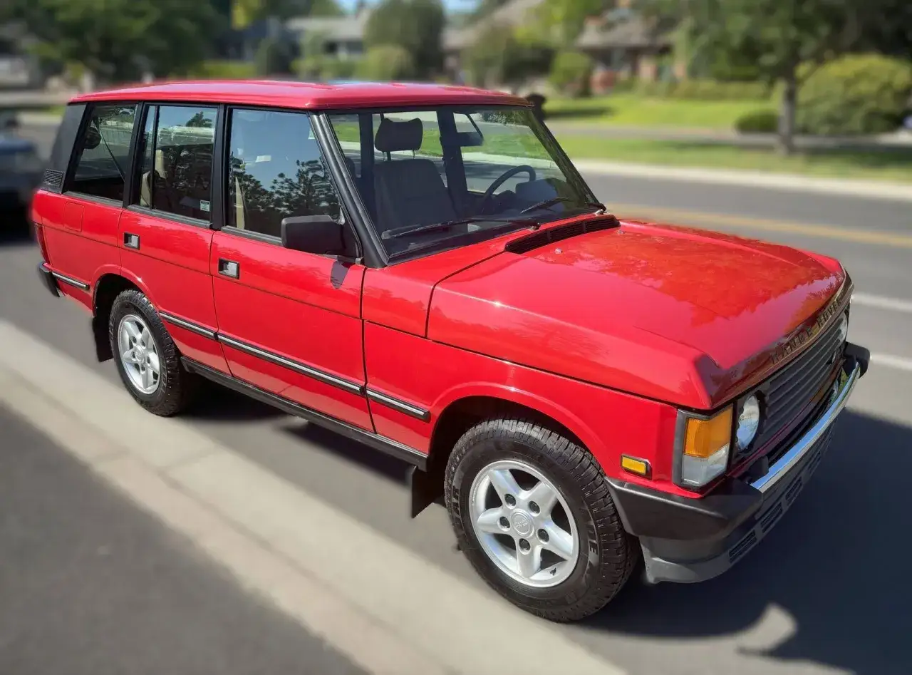 Ein klassischer, knallroter Range Rover steht am Straßenrand. Sein glänzender Lack reflektiert die Sonne, während die markanten Linien seine zeitlose Eleganz unterstreichen.