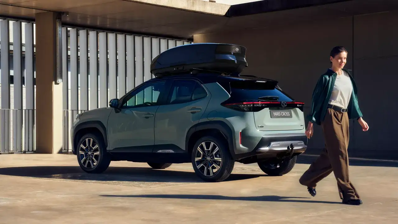 Ein stilvoller Yaris Cross 2026 mit Dachbox, bereit für Abenteuer. Eine Frau geht lässig daran vorbei.