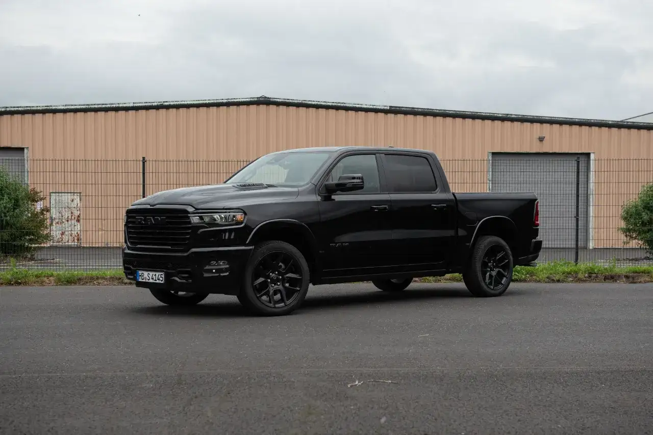 Schwarzer Dodge Ram 1500 Pickup mit dunklen Felgen. Das Fahrzeug steht vor einer Lagerhalle. Das Dodge Ram 1500 Gewicht ist beeindruckend.