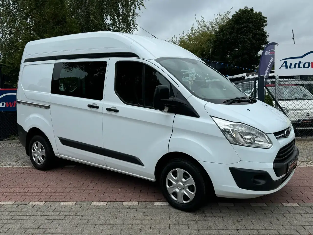 Weißer Ford Transit Custom mit Hochdach, der auf einem gepflasterten Hof steht. Mögliche Ford Transit Custom Probleme sind hier nicht sichtbar.