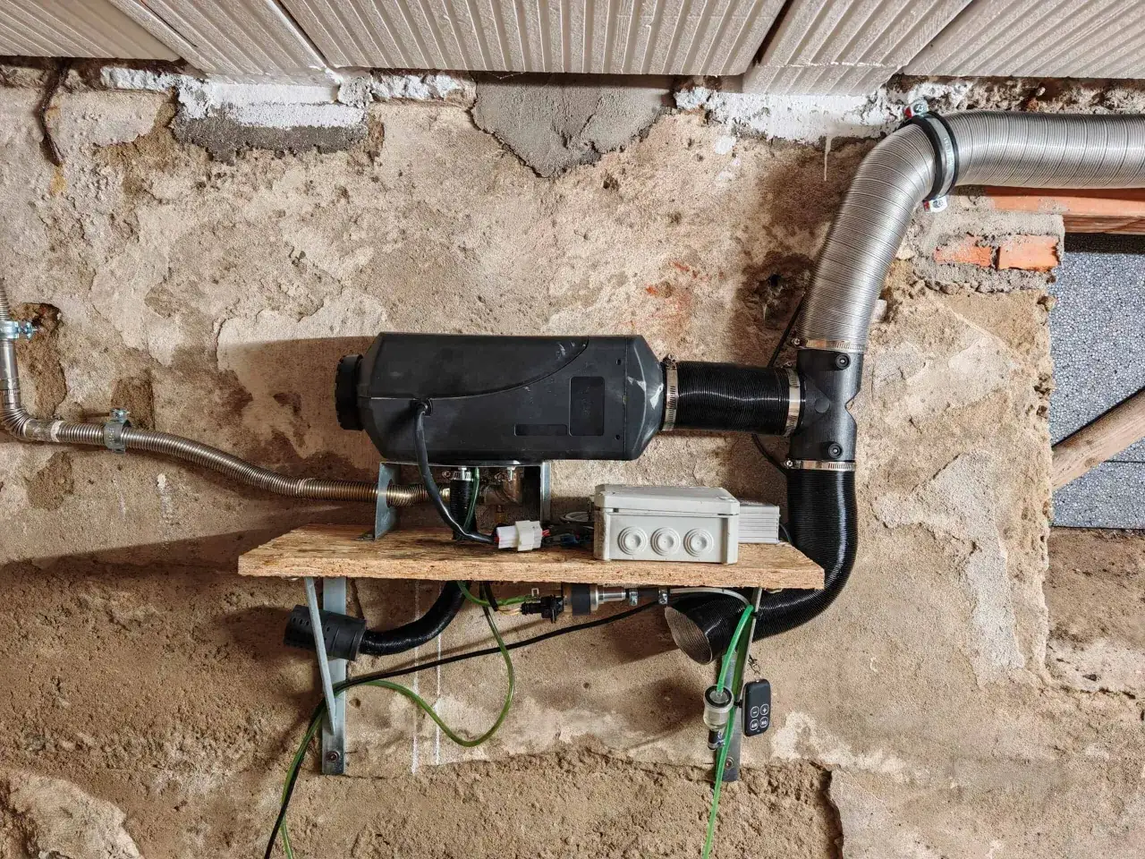 Standheizung einbauen lassen: Schwarzer Heizk&ouml;rper mit flexiblen Rohren und Steuerkasten an einer rauen Wand montiert.
