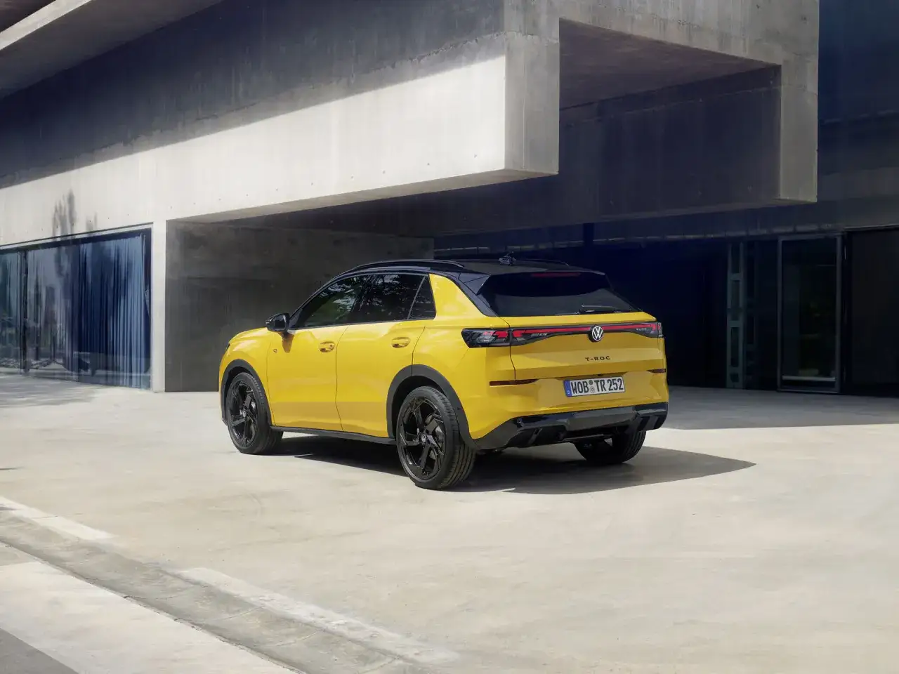 Gelber VW T-Roc mit schwarzem Dach und Felgen parkt vor moderner Betonarchitektur.