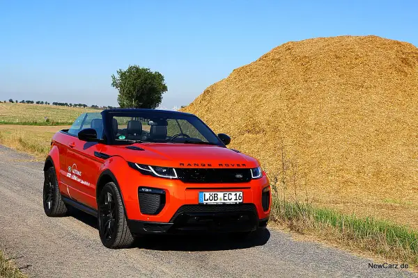 Ein leuchtend orangefarbener Range Rover Evoque Cabrio steht auf einem Feldweg neben einem riesigen Strohballen.
